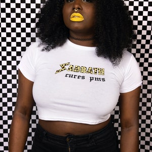 May include: A white crop top with a black and yellow leopard print graphic that says "Sabbath cures pms". The model is wearing black jeans and has yellow lipstick.