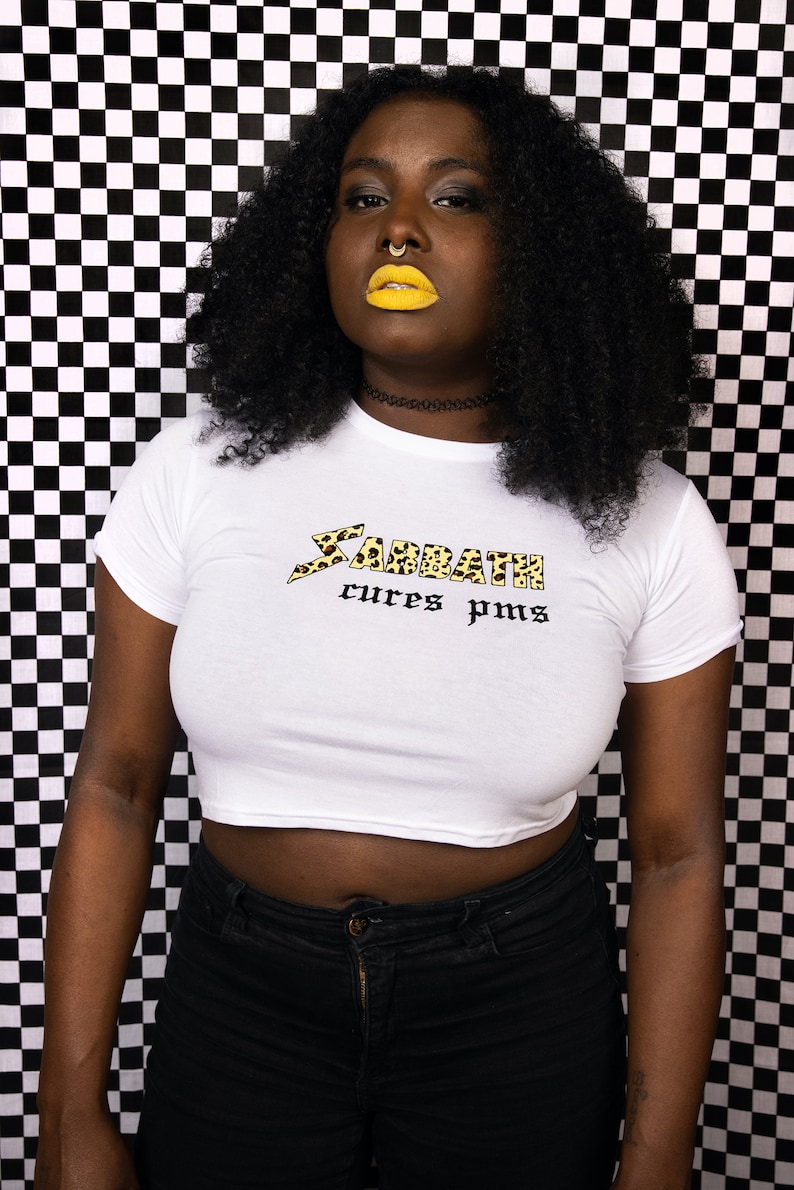 May include: A white crop top with a black and yellow leopard print graphic that says "Sabbath cures pms". The model is wearing black jeans and has yellow lipstick.