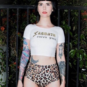 May include: A woman wearing a white crop top with the text "Sabbath cures pms" in gold lettering. She is also wearing leopard print high-waisted shorts. She has tattoos on both arms.
