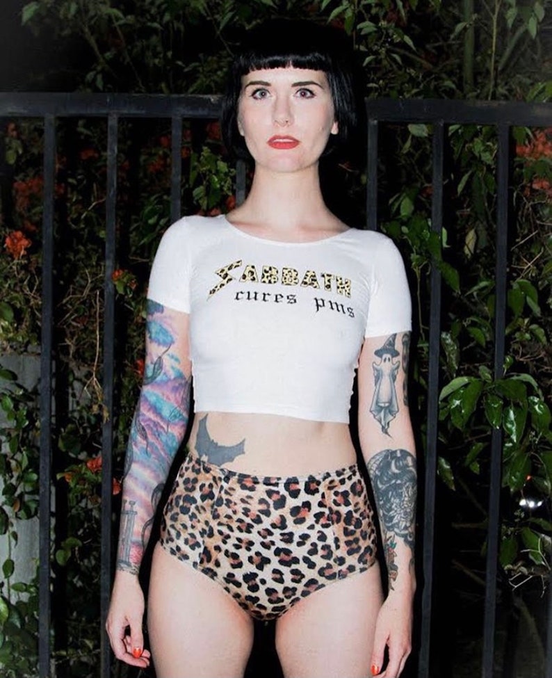 May include: A woman wearing a white crop top with the text "Sabbath cures pms" in gold lettering. She is also wearing leopard print high-waisted shorts. She has tattoos on both arms.
