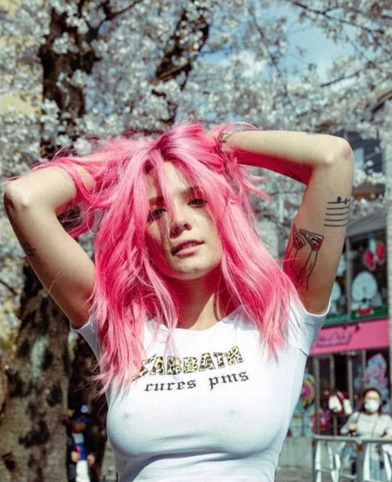 May include: A woman with pink hair is wearing a white crop top with the text "Sabbath cures PMS" printed on it. The text is in a black font with a leopard print pattern.