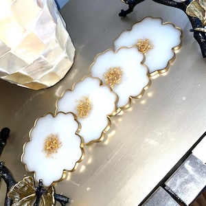 May include: A set of four white resin coasters with a gold rim and gold flecks in the center. The coasters have a scalloped edge and are arranged in a row on a gold surface. Decorative home decor.