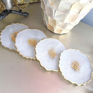 May include: Four white resin coasters with gold edges and gold flecks in the center. The coasters have a wavy edge design. The coasters are on a light-colored surface. A decorative vase is in the background.