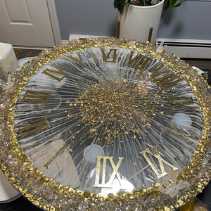 May include: A large, round clock face with Roman numerals and a gold glitter design. The clock face is surrounded by a border of clear crystals and gold paint.