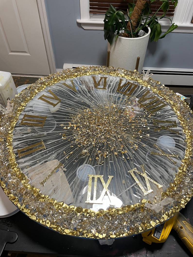 May include: A large, round clock face with Roman numerals and a gold glitter design. The clock face is surrounded by a border of clear crystals and gold paint.
