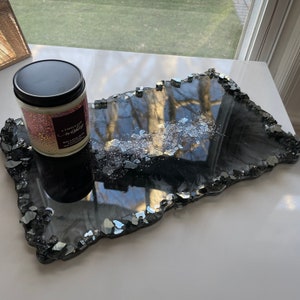 May include: Black resin tray with a jagged edge and silver glitter embedded in the resin. The tray is on a white surface with a candle on top of it. The candle is white with a black lid and has the text "A Thousand Wishes" on the label.