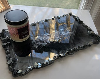 May include: Black resin tray with a jagged edge and silver glitter embedded in the resin. The tray is on a white surface with a candle on top of it. The candle is white with a black lid and has the text "A Thousand Wishes" on the label.