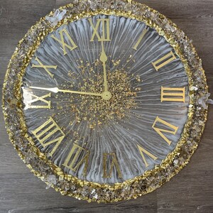 May include: A white and gold resin clock with Roman numeral markings and a gold glitter background. The clock is surrounded by a border of clear and gold crystals.