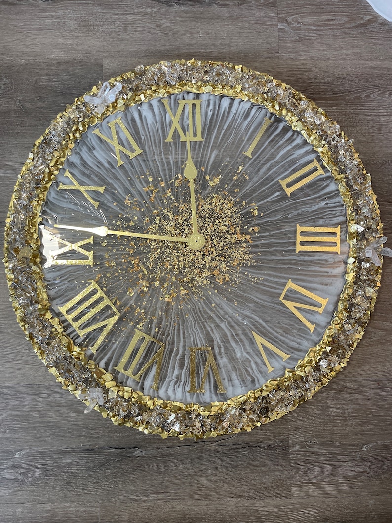 May include: A white and gold resin clock with Roman numeral markings and a gold glitter background. The clock is surrounded by a border of clear and gold crystals.