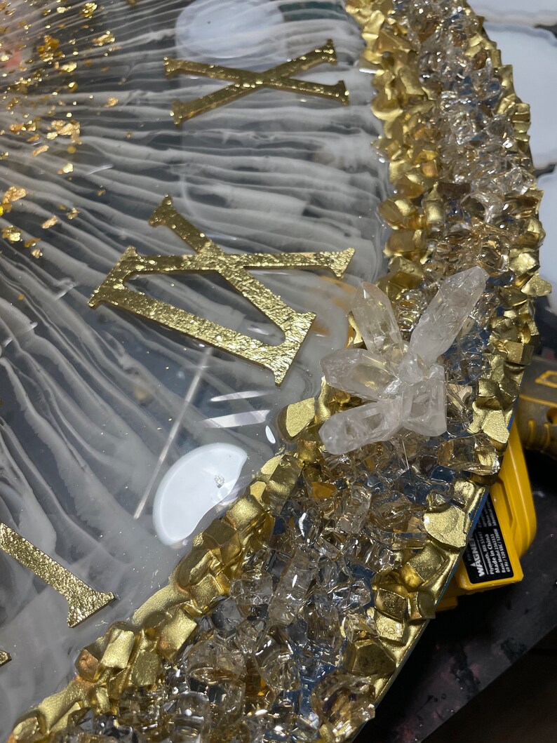 May include: A close-up of a clock face with Roman numerals, gold leaf, and clear quartz crystals. The clock face is set in a white resin base with a gold trim.