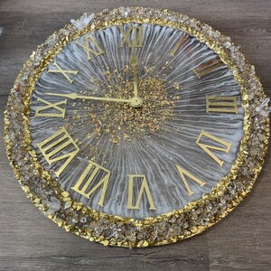 May include: A white and gold resin clock with Roman numeral hour markers and a gold glitter background. The clock is surrounded by a border of clear and gold crystals.