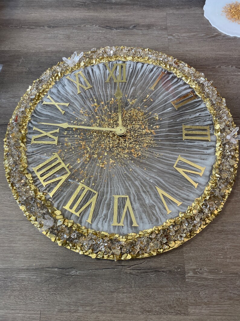 May include: A white and gold resin clock with Roman numeral hour markers and a gold glitter background. The clock is surrounded by a border of clear and gold crystals.