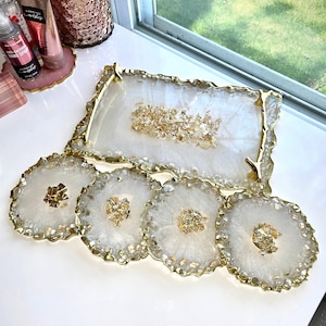 Könnte beinhalten: Ein weißes Resin-Tablett und ein passendes Untersetzer-Set mit Goldblatt-Akzenten. Das Tablett hat eine rechteckige Form mit goldfarbenem Rand, und die Untersetzer sind rund mit goldenen Rändern. Das Set ist für dekorative Zwecke bestimmt.