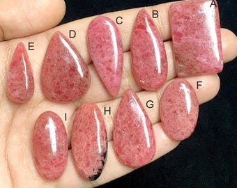 May include: A collection of nine polished rhodochrosite gemstones in various shapes, including teardrop, oval, and rectangular. The stones display a range of pink hues with natural veining and patterns. The gemstones are displayed against a dark background.