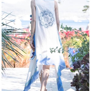 May include: A white sleeveless tunic with blue and white stripes and a blue mandala design on the back. The tunic has a long, flowing hemline and is decorated with colorful pom-poms.