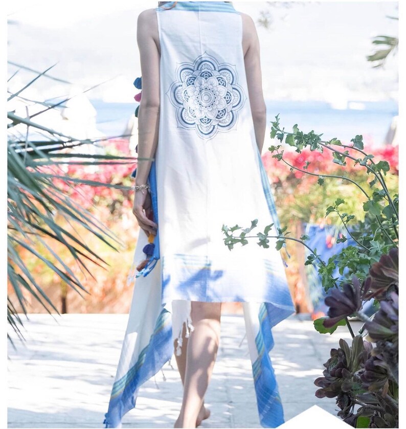May include: A white sleeveless tunic with blue and white stripes and a blue mandala design on the back. The tunic has a long, flowing hemline and is decorated with colorful pom-poms.