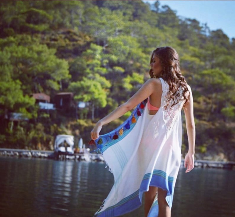 May include: A woman wearing a white crochet top with a blue and white striped cover-up. The cover-up has colorful tassels and is worn over a pink bikini top. The woman is standing on a dock with a body of water behind her.
