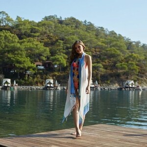 May include: A woman wearing a white and blue striped beach cover-up with pom poms on the fringe. She is standing on a wooden dock with a body of water and a green hillside in the background.