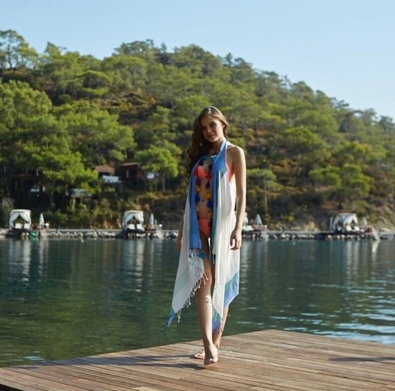 May include: A woman wearing a white and blue striped beach cover-up with pom poms on the fringe. She is standing on a wooden dock with a body of water and a green hillside in the background.