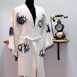 May include: White cotton robe with blue and black eye designs. The robe has a tie belt and fringe trim.