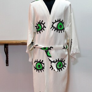 May include: White cotton robe with a green and black eye print. The robe has a tie belt and fringe trim.