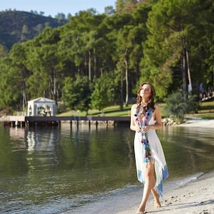 May include: A woman in a white dress with blue and red trim walks on a sandy beach. The beach is near a body of water with a wooden dock and a gazebo in the background.
