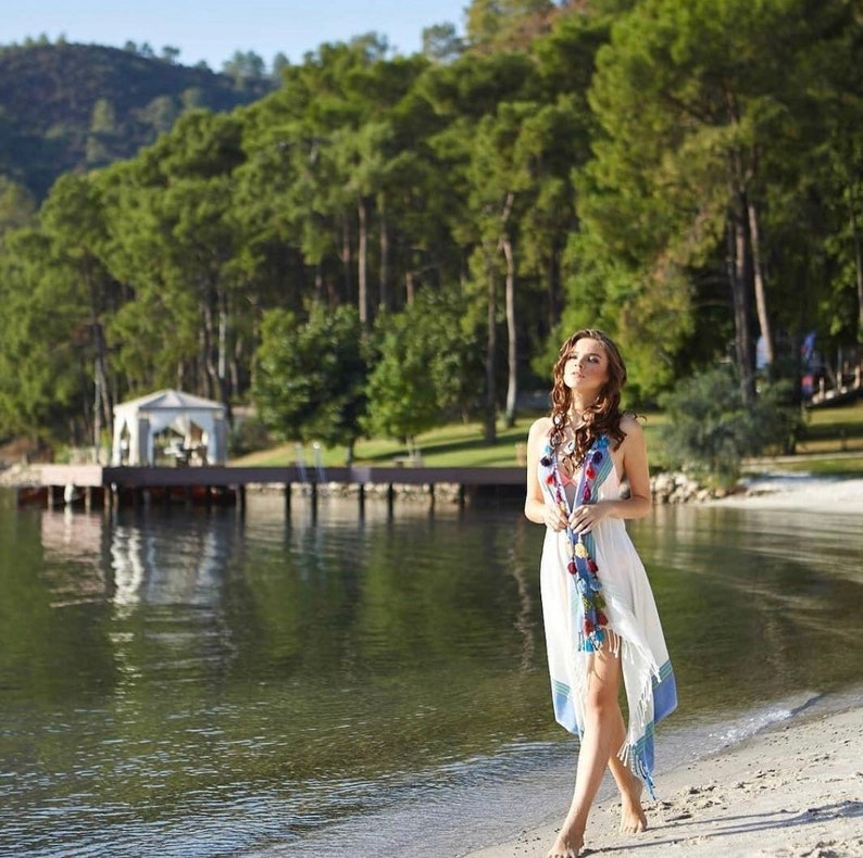 May include: A woman in a white dress with blue and red trim walks on a sandy beach. The beach is near a body of water with a wooden dock and a gazebo in the background.