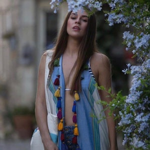 May include: A woman wearing a blue and white patterned vest with colorful tassels. The vest has a white and blue striped pattern and is open in the front, revealing a white bikini top. The woman is standing in front of a bush with blue flowers.