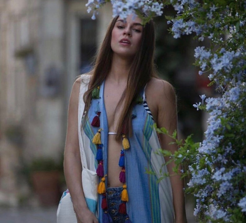 May include: A woman wearing a blue and white patterned vest with colorful tassels. The vest has a white and blue striped pattern and is open in the front, revealing a white bikini top. The woman is standing in front of a bush with blue flowers.