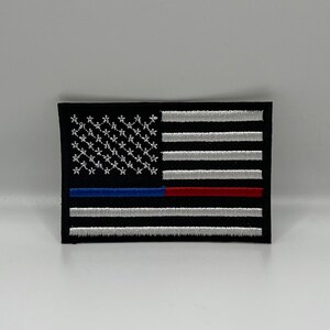 May include: Embroidered patch featuring a stylized American flag design. The flag has a black background with white stars, white and gray stripes, and blue and red stripes. The patch is rectangular and has a black border.