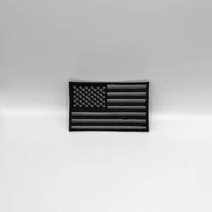 May include: A black and gray embroidered patch featuring the American flag design. The flag has gray stripes and a field of gray stars. The patch is rectangular and has a black border.