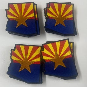 May include: A collection of embroidered patches in the shape of the state of Arizona. Each patch features the Arizona state flag design with a blue lower half, a yellow star, and a red and yellow sunburst.