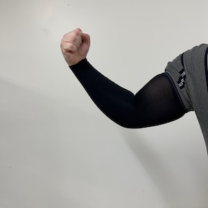 May include: Black compression sleeve worn on the arm. The sleeve covers the arm from the wrist to the upper arm. The person is flexing their arm, showing the sleeve's fit and coverage. The background is a plain white wall.