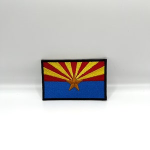May include: Embroidered patch featuring the Arizona state flag. The design includes a red and yellow sunburst over a blue field, with a copper star in the center. The patch has a black border.