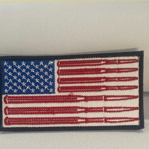 May include: Embroidered patch featuring the American flag design, with bullets replacing the stripes. The flag's blue field has white stars, and the stripes are red bullets. The patch has a black border.