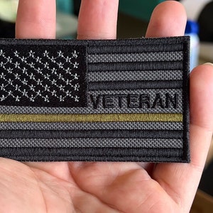 May include: A rectangular patch featuring a stylized American flag design. The flag has a black and gray color scheme with white embroidered stars and the word "VETERAN" in white. A gold line runs horizontally across the center.