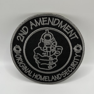May include: A round black patch with gray embroidered text and a handgun graphic. The text reads "2ND AMENDMENT" and "ORIGINAL HOMELAND SECURITY". The patch has a gray border and bullet hole details.