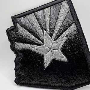 May include: Embroidered patch in the shape of Arizona, featuring a gray star and radiating lines against a black background. The patch has a textured appearance and a black border.