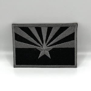 May include: A rectangular patch featuring the Arizona state flag design. The top half displays gray sun rays emanating from a central star on a black background. The bottom half is solid black, with a gray border.
