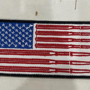 May include: Embroidered patch featuring a stylized American flag design. The blue field has white stars, and the red stripes are replaced with a series of rifle barrels. The patch has a black border.