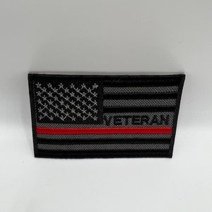 May include: A rectangular patch featuring a stylized American flag design. The flag is primarily black and gray, with a red stripe and the word "VETERAN" in white. The stars are white.