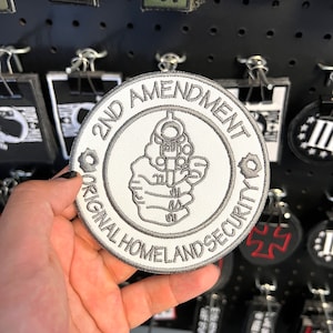 May include: A round, white patch with gray embroidered text and a gun graphic. The text reads "2ND AMENDMENT ORIGINAL HOMELAND SECURITY". The patch is held up by a hand, with other patches visible in the background.