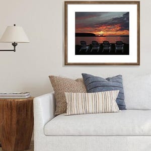 May include: A framed photograph of a sunset over a lake with five chairs facing the water. The sky is a vibrant orange and pink with dark clouds. The chairs are silhouetted against the water.