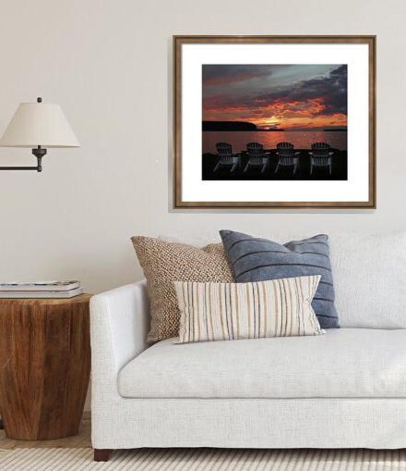 May include: A framed photograph of a sunset over a lake with five chairs facing the water. The sky is a vibrant orange and pink with dark clouds. The chairs are silhouetted against the water.