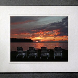 May include: Four white chairs facing a lake with a colorful sunset in the background.