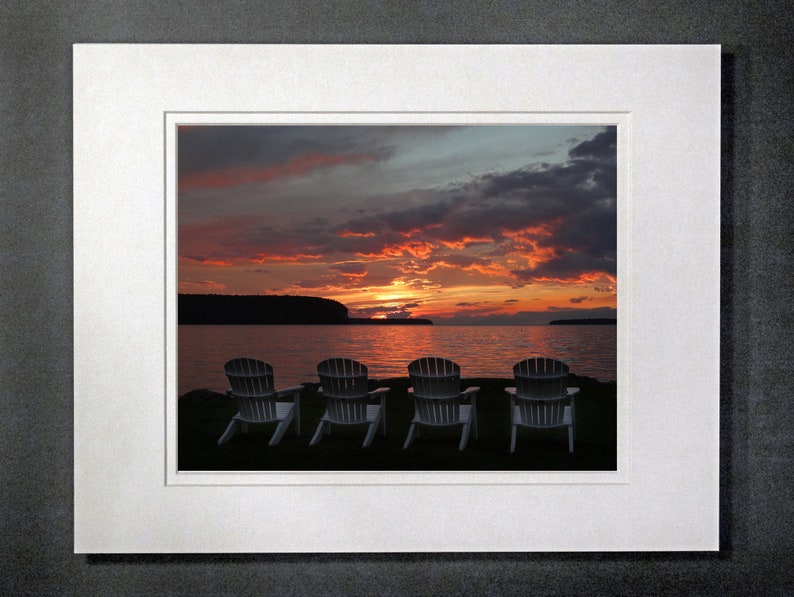 May include: Four white chairs facing a lake with a colorful sunset in the background.