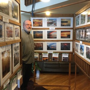 May include: A man stands in front of a display of framed photographs of landscapes. The photos are arranged on wooden shelves and feature a variety of scenes, including sunsets, forests, and bodies of water. The man is wearing a green shirt and has a beard.