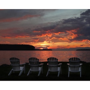 May include: Four white wooden Adirondack chairs facing a lake with a vibrant sunset in the background.