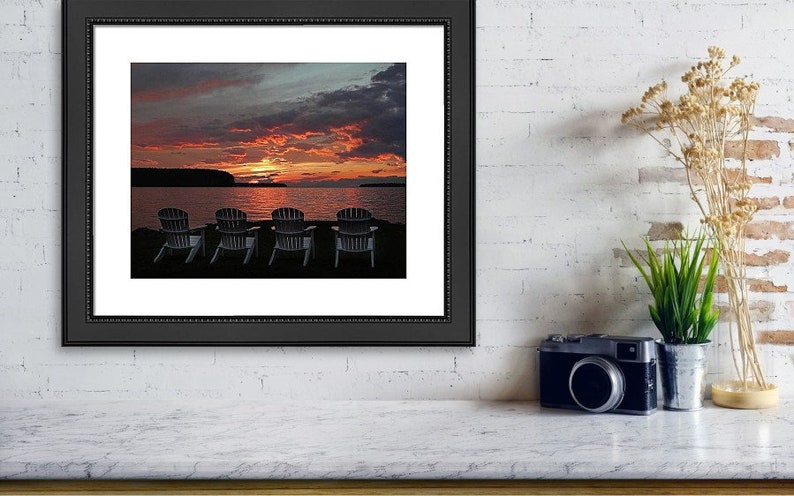 May include: Four Adirondack chairs face a lake at sunset. The sky is a vibrant mix of orange, pink, and purple with dark clouds. The water is calm and reflects the colors of the sky.