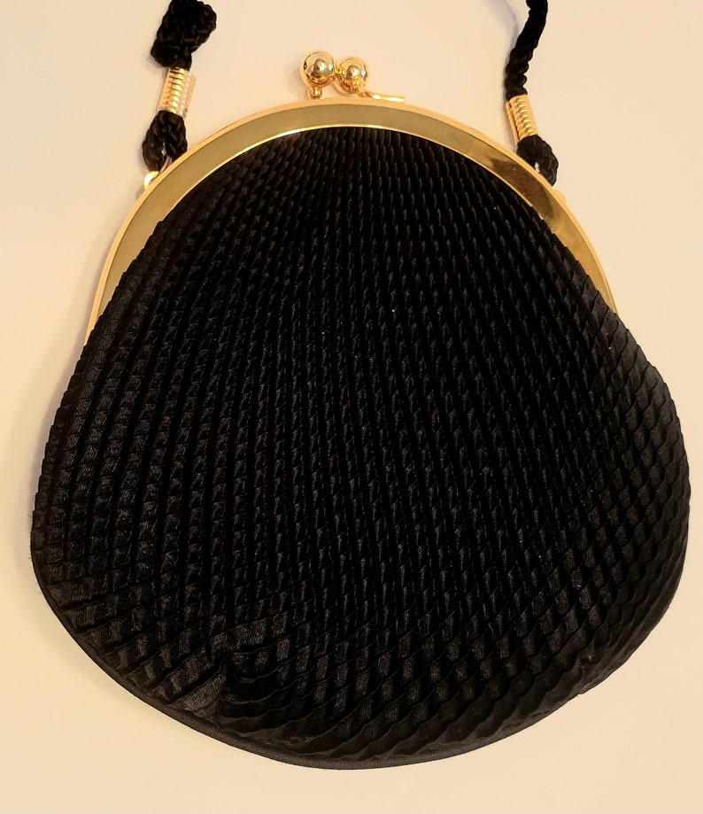 May include: A black, vintage-style purse with a gold-colored frame and clasp. The purse has a textured, pleated design and a black cord handle. The clasp features two small, gold-colored spheres.